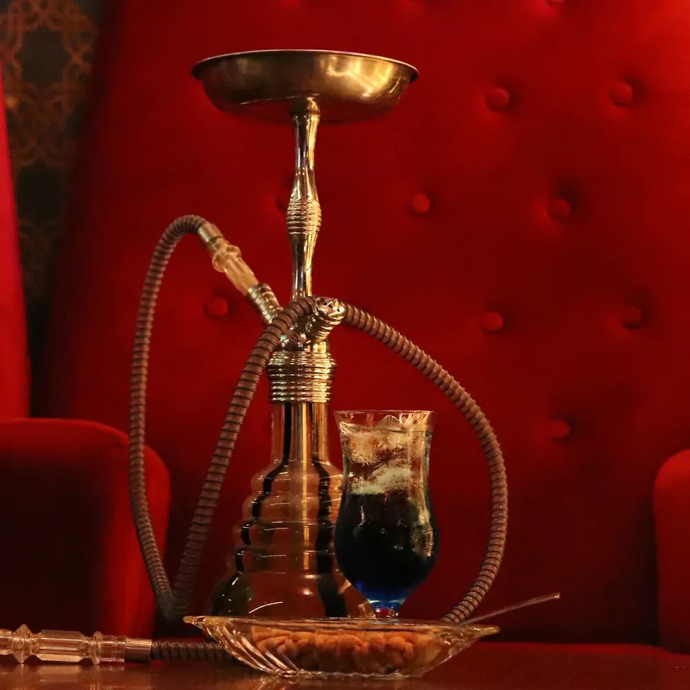 A brass hookah with a coiled hose on a glass base, in front of a red seat